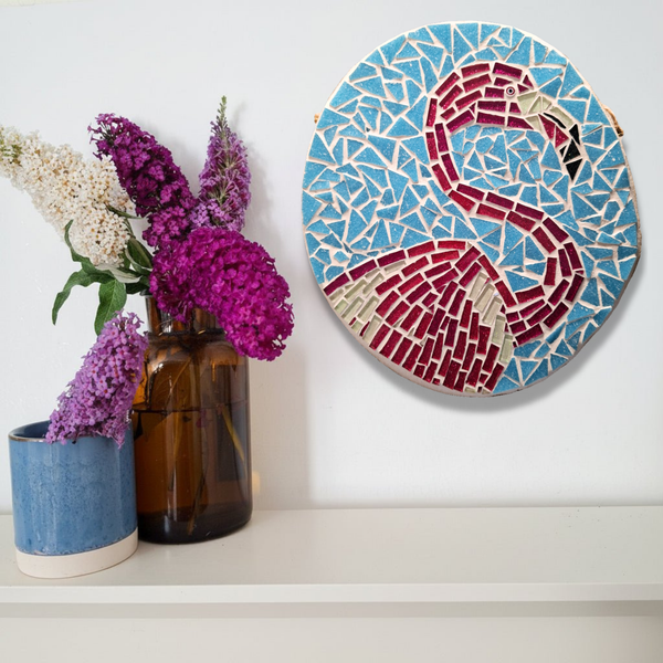 A round mosaic wall art piece featuring a flamingo design in red, cream, and light blue, displayed on a white wall above a shelf with decorative vases and flowers.