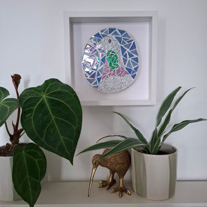 A square white-framed mosaic wall art piece featuring a stylized pigeon in blue, silver, green, and pink glass tiles, displayed on a shelf above potted plants and a small gold kiwi figurine.
