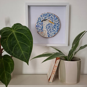 A framed circular mosaic art piece featuring a white heron with a yellow beak on a blue and white geometric background, displayed on a shelf next to green potted plants.