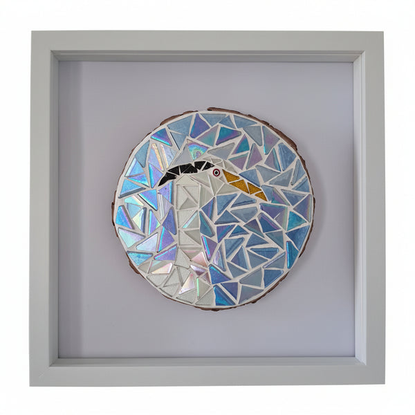 A framed mosaic art piece depicting a heron in shades of blue, white, and silver, mounted on a light gray background inside a white square frame.