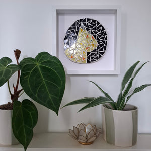 A circular mosaic wall art piece with a black and white geometric border and a central gold-toned abstract cat design, displayed in a white square frame on a wall above a shelf with potted plants.