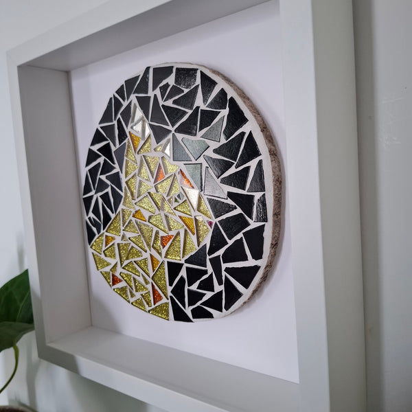 A circular mosaic wall art piece with a gold cat design mounted inside a white square shadow box frame.