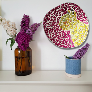 A ginger cat mosaic styled on a white wall above a shelf with purple flowers in an amber glass vase and a blue ceramic cup.