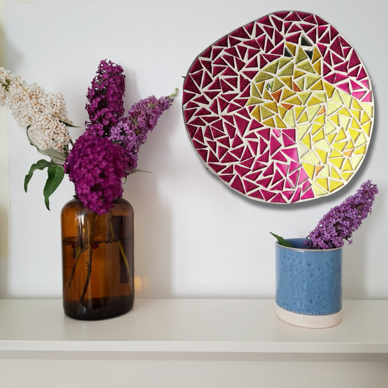 A ginger cat mosaic styled on a white wall above a shelf with purple flowers in an amber glass vase and a blue ceramic cup.