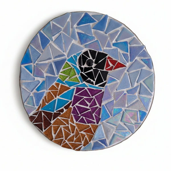 Circular mosaic wall art featuring a stylized bird design. Crafted with colourful glittery glass pieces on an iridescent blue background, set on rustic wood.
