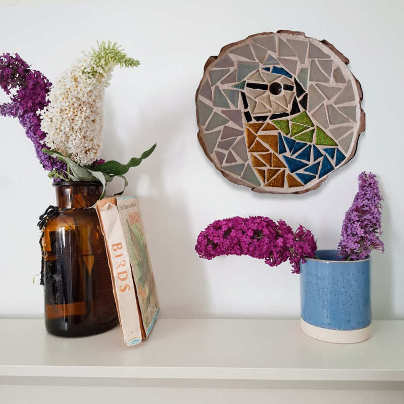 A styled interior shelf scene with dried flowers in an amber glass bottle and a blue ceramic pot, next to a round mosaic wall hanging featuring a great tit bird.