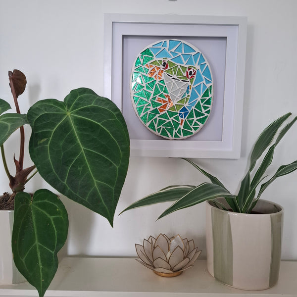 A square white-framed mosaic wall art piece featuring a colorful frog design hangs on a white wall above a shelf with potted plants.