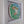 A framed mosaic art piece depicting a frog, mounted on a white backing and enclosed in a white square frame, shown against a plain wall.