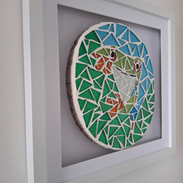A framed mosaic art piece depicting a frog, mounted on a white backing and enclosed in a white square frame, shown against a plain wall.