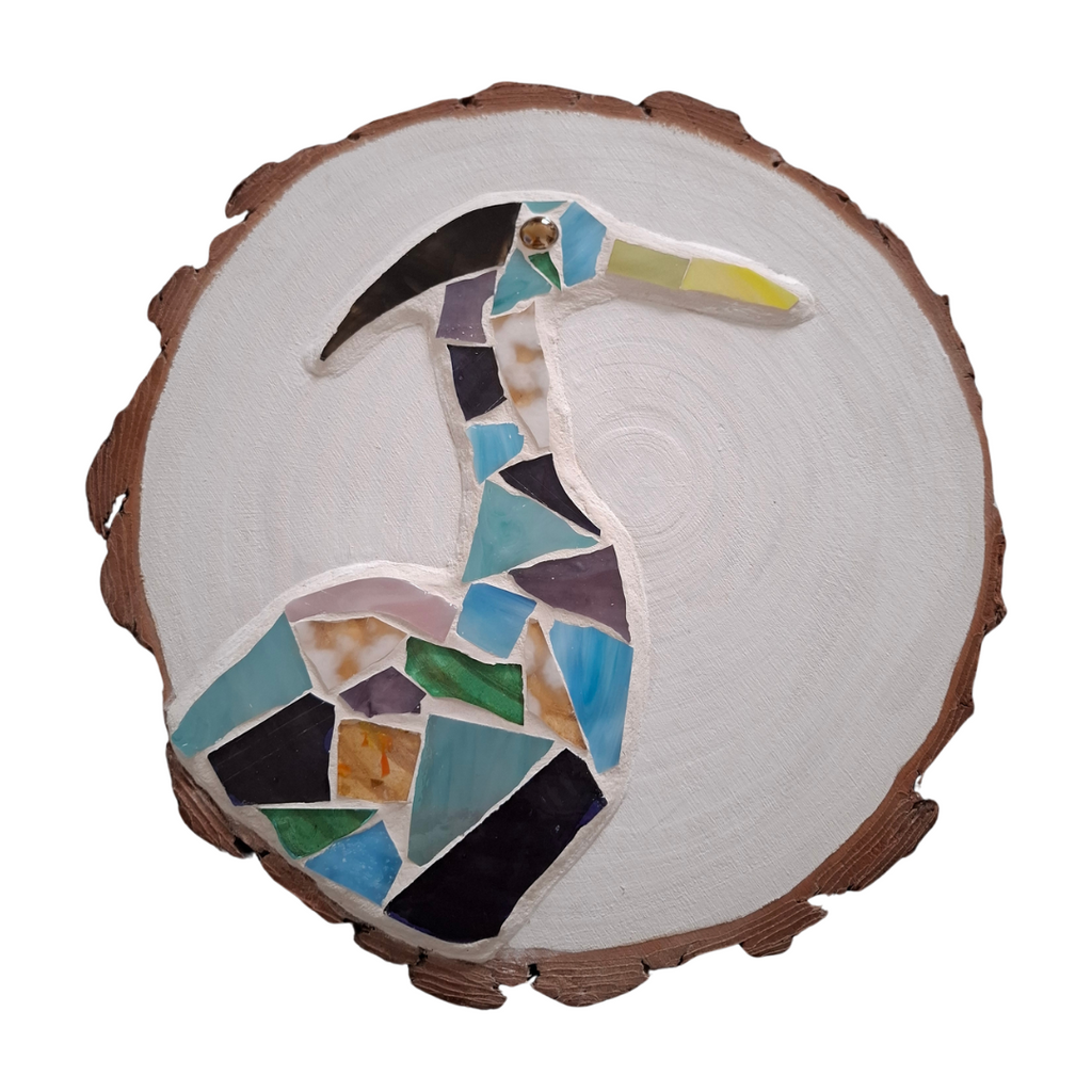 Circular mosaic wall art featuring a stylized heron design. Crafted with blue, green and aqua stained glass pieces on a striking white painted background, set on rustic wood.