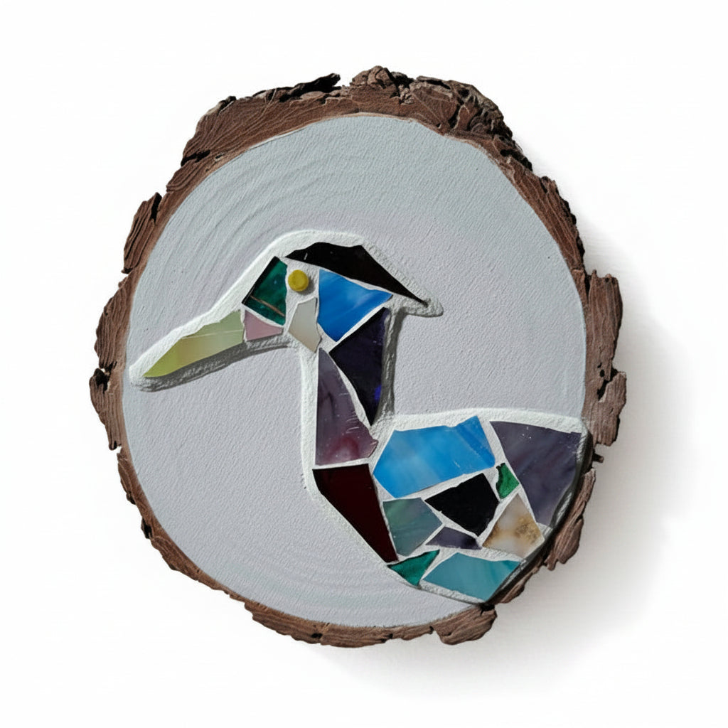 Circular mosaic wall art featuring a stylized heron design. Crafted with blue, green and aqua stained glass pieces on a striking white painted background, set on rustic wood.