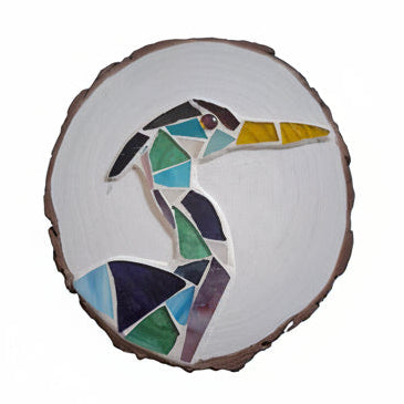 Circular mosaic wall art featuring a stylized heron design. Crafted with blue, green and aqua stained glass pieces on a striking white painted background, set on rustic wood.