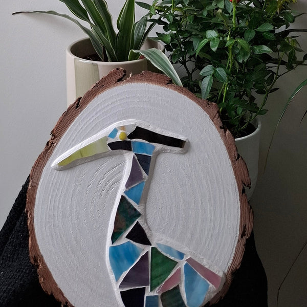 Circular mosaic wall art featuring a stylized heron design. Crafted with blue, green and aqua stained glass pieces on a striking white painted background, set on rustic wood.