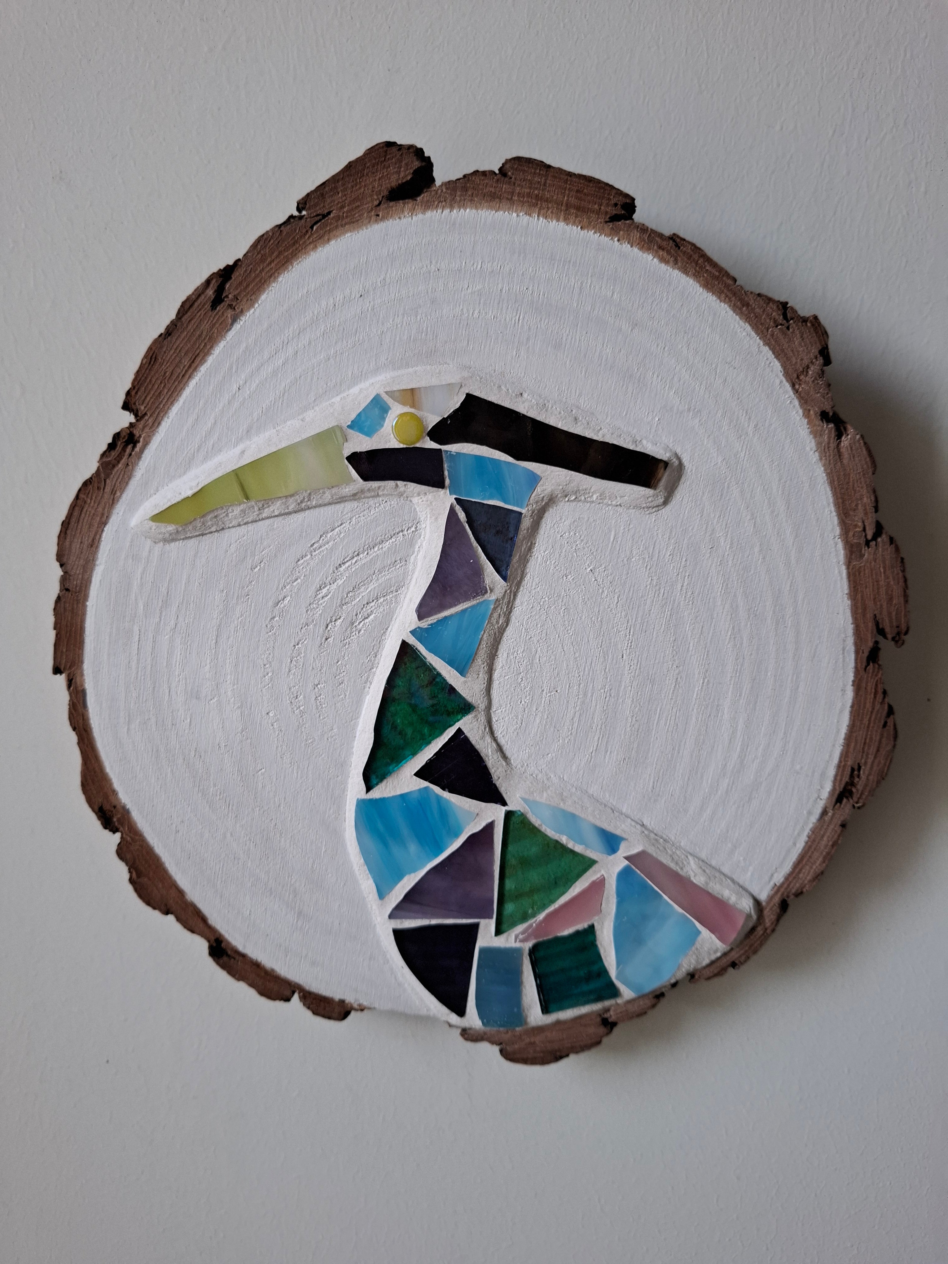 Circular mosaic wall art featuring a stylized heron design. Crafted with blue, green and aqua stained glass pieces on a striking white painted background, set on rustic wood.