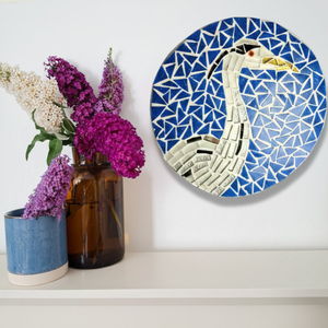 A round mosaic wall art piece featuring a heron design in blue, white, black, and silver, displayed on a white wall above a shelf with decorative vases and flowers.