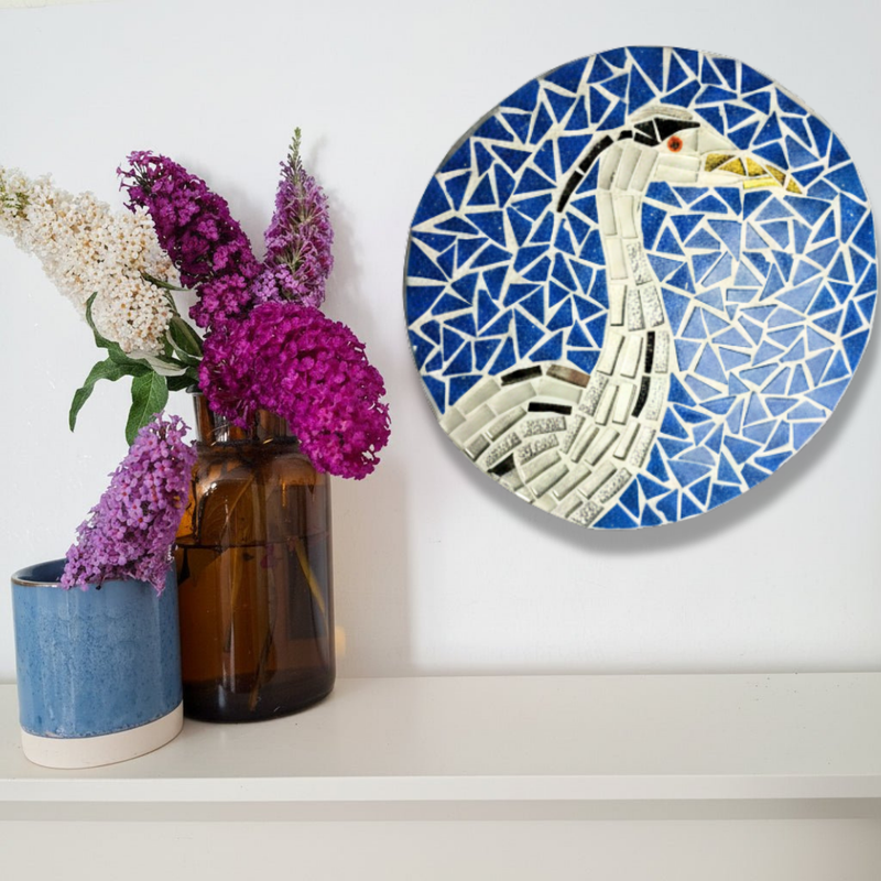 A round mosaic wall art piece featuring a heron design in blue, white, black, and silver, displayed on a white wall above a shelf with decorative vases and flowers.