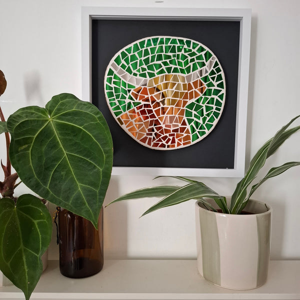 Framed mosaic wall art featuring a highland cow in brown, bronze and gold toned reflective mirror tiles on a green background. The mosaics sits above a shelf with potted plants. 