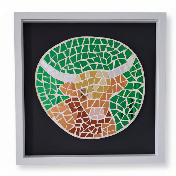 A square framed mosaic art piece featuring a stylized Highland cow head. The cow is composed of green, orange, brown, and bronze mosaic tiles with silver horns, enclosed in a white frame, mounted on a light gray wall.