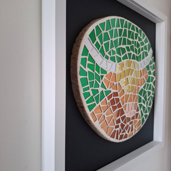 A circular wood slice mosaic wall art piece featuring a stylized highland cow, mounted on a black backing within a white frame.