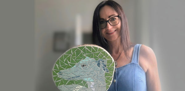 Artist Naomi Hope holds a round mosaic featuring a greyhound in green and silver tiles. She is wearing a light blue denim top against a softly blurred indoor background.