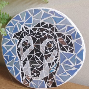 A circular mosaic wall art piece featuring a stylized dog made of blue, white, black, and brown glass tiles, leaning on a wooden surface next to a green fern plant.