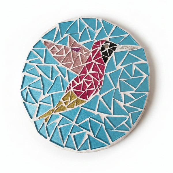 Circular mosaic wall art featuring a stylized hummingbird design. Crafted with glittery pink and gold glass pieces on a sparkly blue background, set on rustic wood.