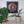 A framed circular mosaic wall art piece featuring a dog with red, black, white, and silver, displayed on a shelf above green potted plants.