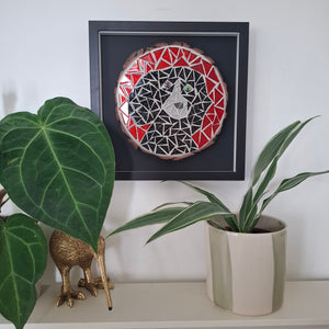 A framed circular mosaic wall art piece featuring a dog with red, black, white, and silver, displayed on a shelf above green potted plants.