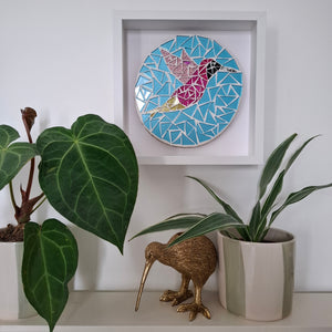 A circular mosaic wall art piece featuring a stylized pink hummingbird, framed, above a shelf with potted plants. 