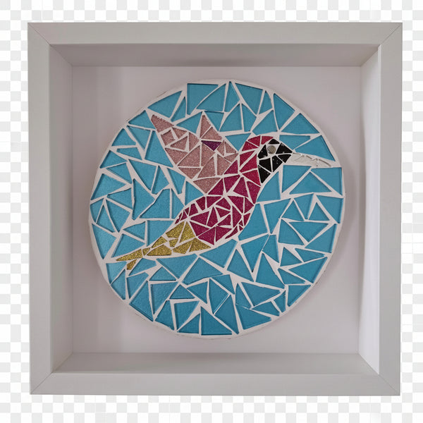 A circular mosaic wall art piece featuring a stylized hummingbird in pink, yellow, and black, set against a blue geometric background and mounted in a white square shadow box frame.