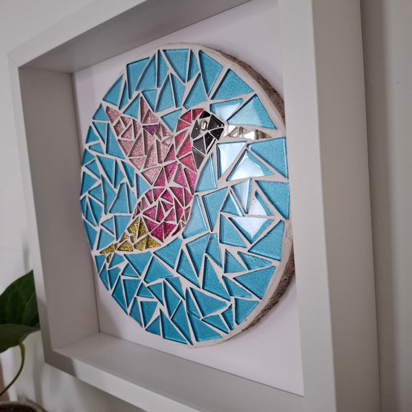 A circular mosaic hummingbird in pink and blue glittery tiles framed on a wall.