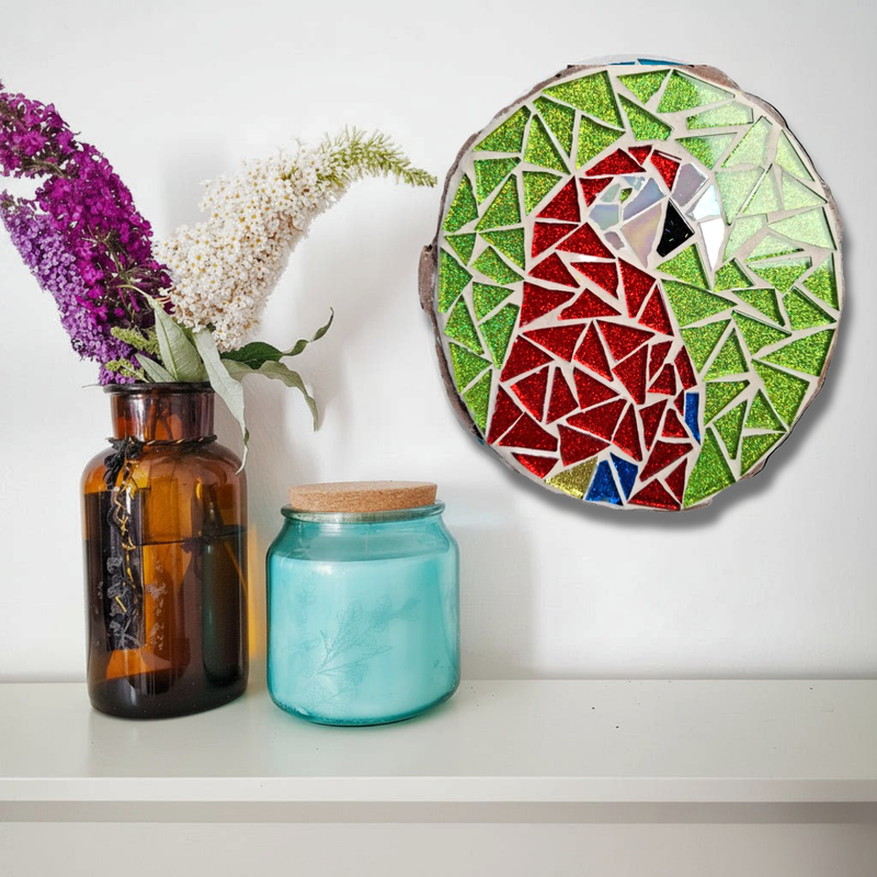 A circular glass mosaic featuring a parrot design, mounted on a white wall above a shelf. The parrot is made of red, green, blue, and white mosaic glass.