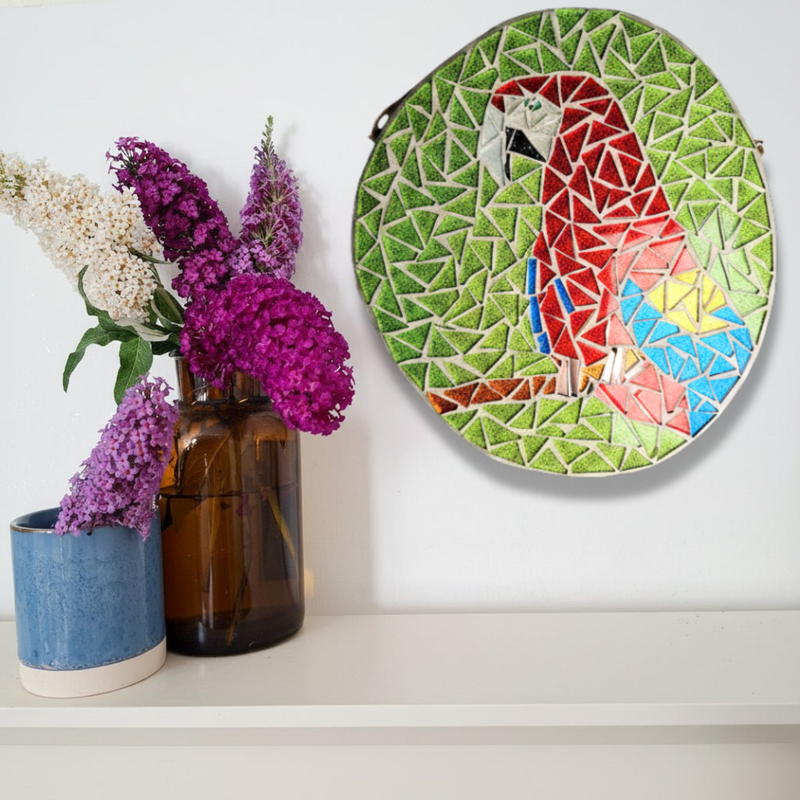 A colorful mosaic featuring a parrot, displayed on a white wall above a shelf with two vases of flowers.