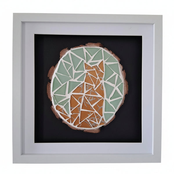 A square framed mosaic art piece featuring a stylized cat design. The artwork is composed of light green, gold, and white mosaic tiles on a dark background, housed in a white square frame.