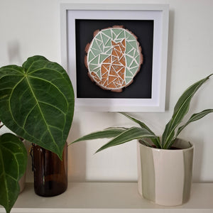 Circular mosaic wall art featuring a golden cat silhouette, framed on a wall above potted plants 