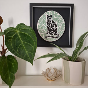 A framed circular mosaic wall art piece featuring a black cat design hangs on a white wall above a shelf with potted plants and a small decorative lotus holder.