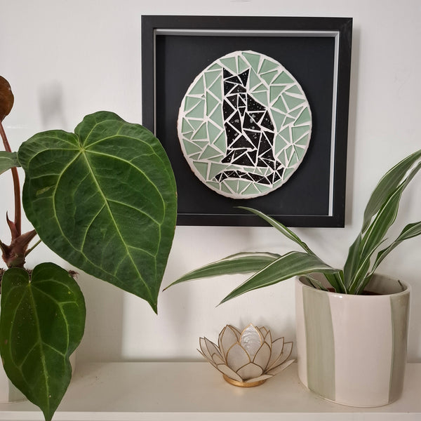 A framed circular mosaic wall art piece featuring a black cat design hangs on a white wall above a shelf with potted plants and a small decorative lotus holder.