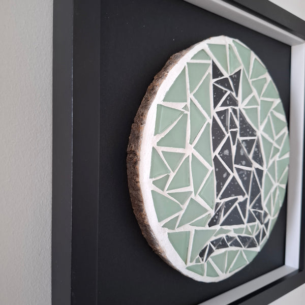 A circular mosaic wall art piece with a natural wood slice edge, featuring a black cat design on a light green and white geometric background, mounted on a dark backing board.