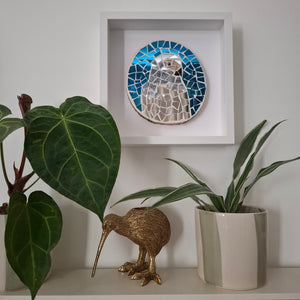 A square white shadow box frame displaying a reflective silver parrot mosaic wall art piece, styled on a shelf with green potted plants and a gold kiwi bird figurine.