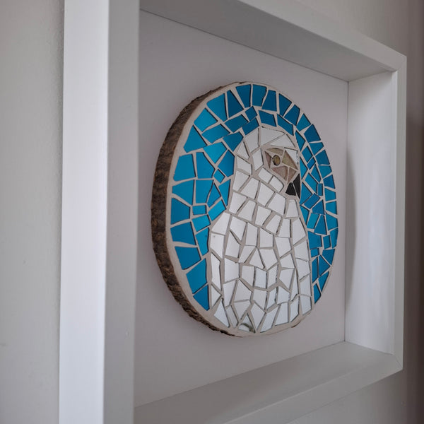 A circular mosaic wall art piece featuring a stylized bird in silver and blue, mounted on a white recessed shelf against a neutral wall.