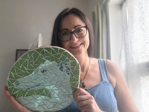 Artist Naomi Hopeholds up a circular mosaic wall hanging featuring a stylized greyhound face made of green, blue, and white tiles.