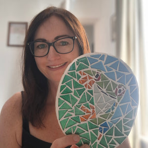 Mosaic artist Naomi Hope holding a round mosaic art piece featuring a frog design in shades of green, blue, orange, and white.