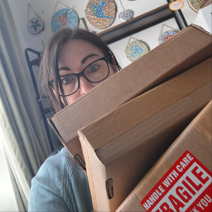 Artist Naomi Hope peeks over 3 large cardboard shipping boxes in a room with decorative wall hangings. The box has a red 'FRAGILE' label on the corner.