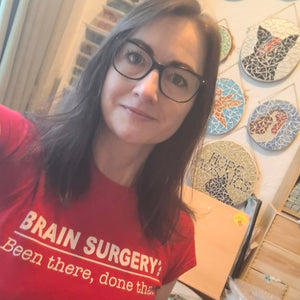 Artist Naomi Hope wearing a red t-shirt with the text 'BRAIN SURGERY: Been there, done that' in white, in a room with mosaic wall art.