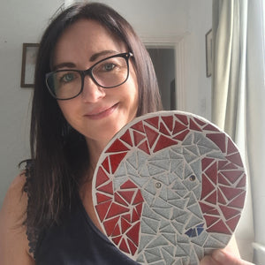 Artist Naomi Hope holds up a mosaic of a silver sparkly greyhound on a red background