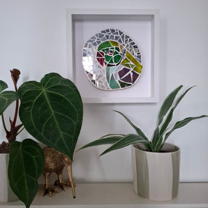 A circular mosaic wall art piece featuring a green parakeet, displayed in a white square shadow box frame on a white wall above a shelf with potted plants.