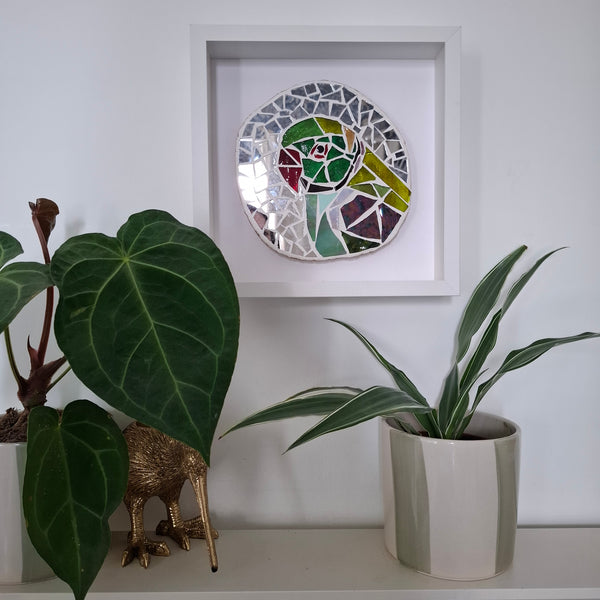 A circular mosaic wall art piece featuring a green parakeet, displayed in a white square shadow box frame on a white wall above a shelf with potted plants.