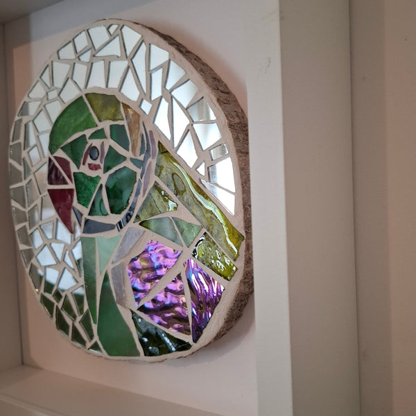 A circular mosaic wall art piece featuring a parakeet design, mounted on a white frame. The mosaic contains green, purple, red, and white glass pieces with a natural wood edge.