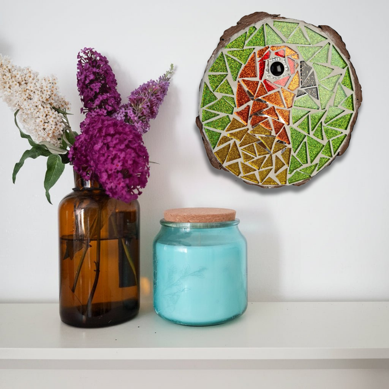 A round mosaic wall art piece featuring a colorful parrot on a green background, displayed on a white wall above a shelf with an amber glass vase of flowers and a blue glass jar.