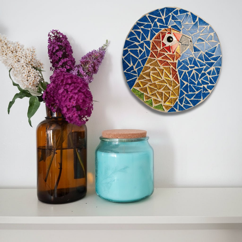 A round mosaic featuring a stylized parakeet in geometric pieces, mounted on a white wall above a shelf. 